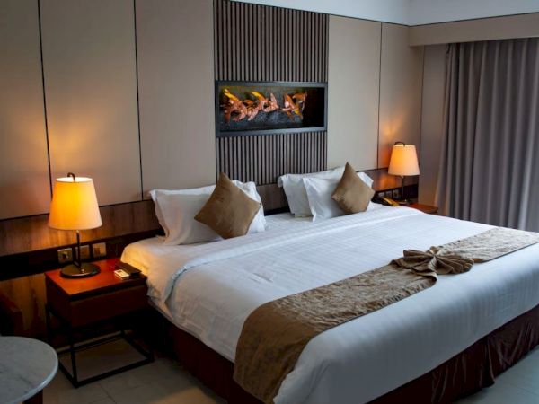A cozy hotel room with a large bed, pillows, two side lamps, a decorative mural above, and a window with closed curtains.