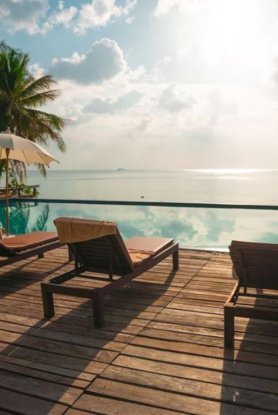 The image shows a serene poolside setup with loungers, umbrellas, and palm trees overlooking a calm sea under a bright sky.