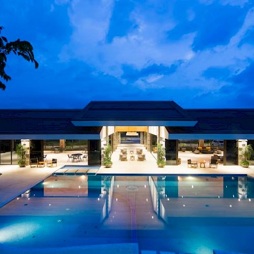 The image shows a luxurious building with a large illuminated pool, surrounded by open spaces and lit interiors, under a twilight sky.