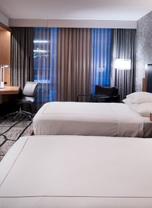 A modern hotel room with two beds, a desk, chair, TV, lamps, and patterned wallpaper, featuring natural lighting from the window.