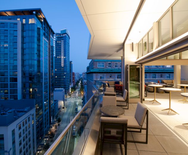 A modern cityscape with tall buildings and a lit-up outdoor balcony featuring tables and chairs in a stylish urban setting.