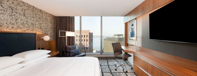 A modern hotel room with a large bed, a flat-screen TV, a desk, and a window offering a city view.
