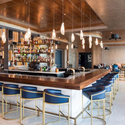 The image shows a modern bar with blue chairs, a well-stocked liquor shelf, and stylish pendant lights. The space looks upscale and inviting.