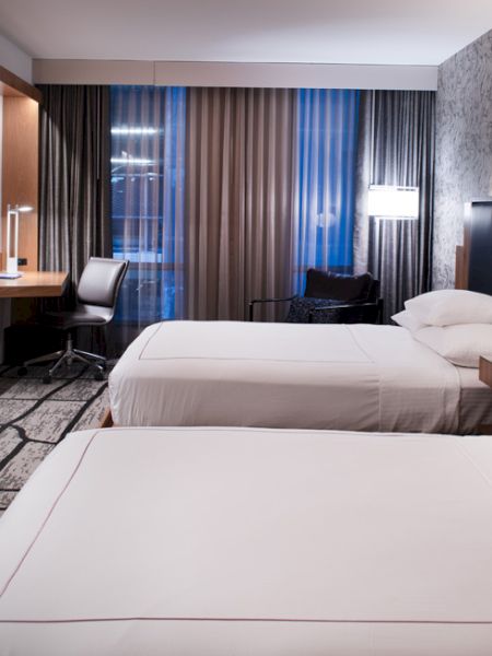 The image shows a modern hotel room with two beds, a TV, desk, and large window with curtains. The walls have a patterned design.