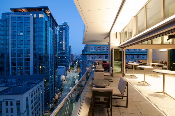 A modern balcony overlooking a cityscape at dusk, with tall buildings and a stylish, partially open indoor seating area.