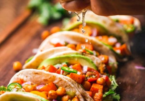 Hand squeezing lime over a row of tacos filled with chickpeas, diced tomatoes, onions, cilantro, and avocado slices on a wooden surface.