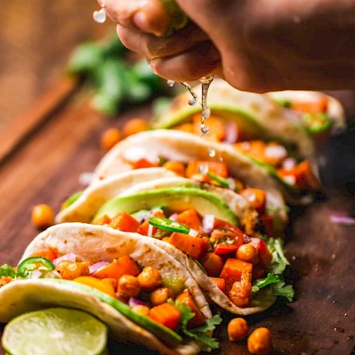 Hand squeezing lime over a row of tacos filled with chickpeas, diced tomatoes, onions, cilantro, and avocado slices on a wooden surface.