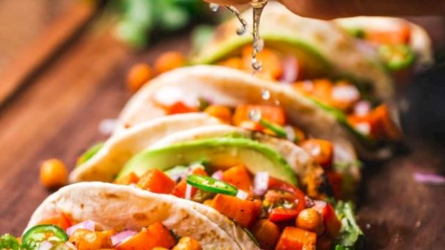 Hand squeezing lime over a row of tacos filled with chickpeas, diced tomatoes, onions, cilantro, and avocado slices on a wooden surface.