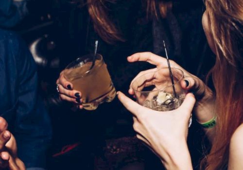 People are enjoying drinks at a social gathering, laughing and engaging in conversation in a dimly lit setting.