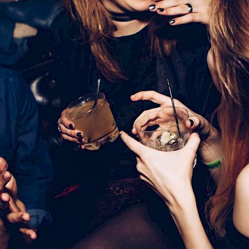 People are enjoying drinks at a social gathering, laughing and engaging in conversation in a dimly lit setting.