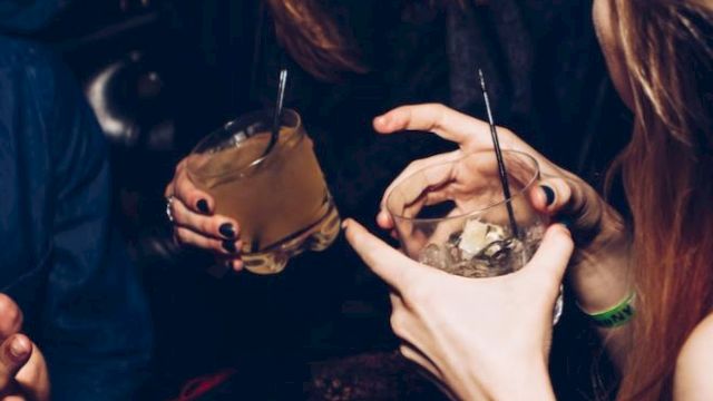 People are enjoying drinks at a social gathering, laughing and engaging in conversation in a dimly lit setting.
