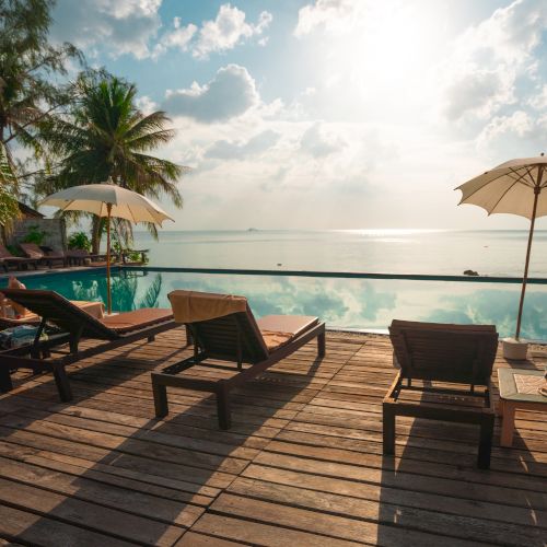 Lounge chairs and umbrellas by an infinity pool overlook a calm sea, with palm trees and a bright sky in the background.