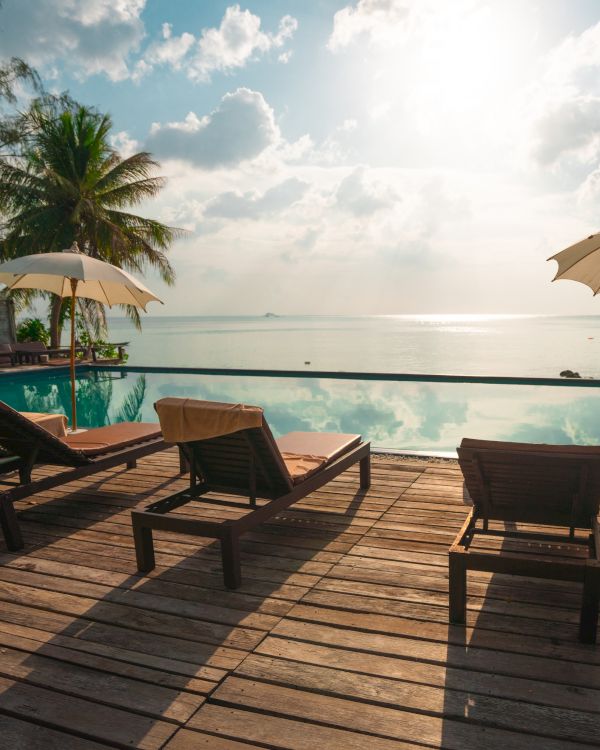 Lounge chairs and umbrellas by an infinity pool overlook a calm sea, with palm trees and a bright sky in the background.