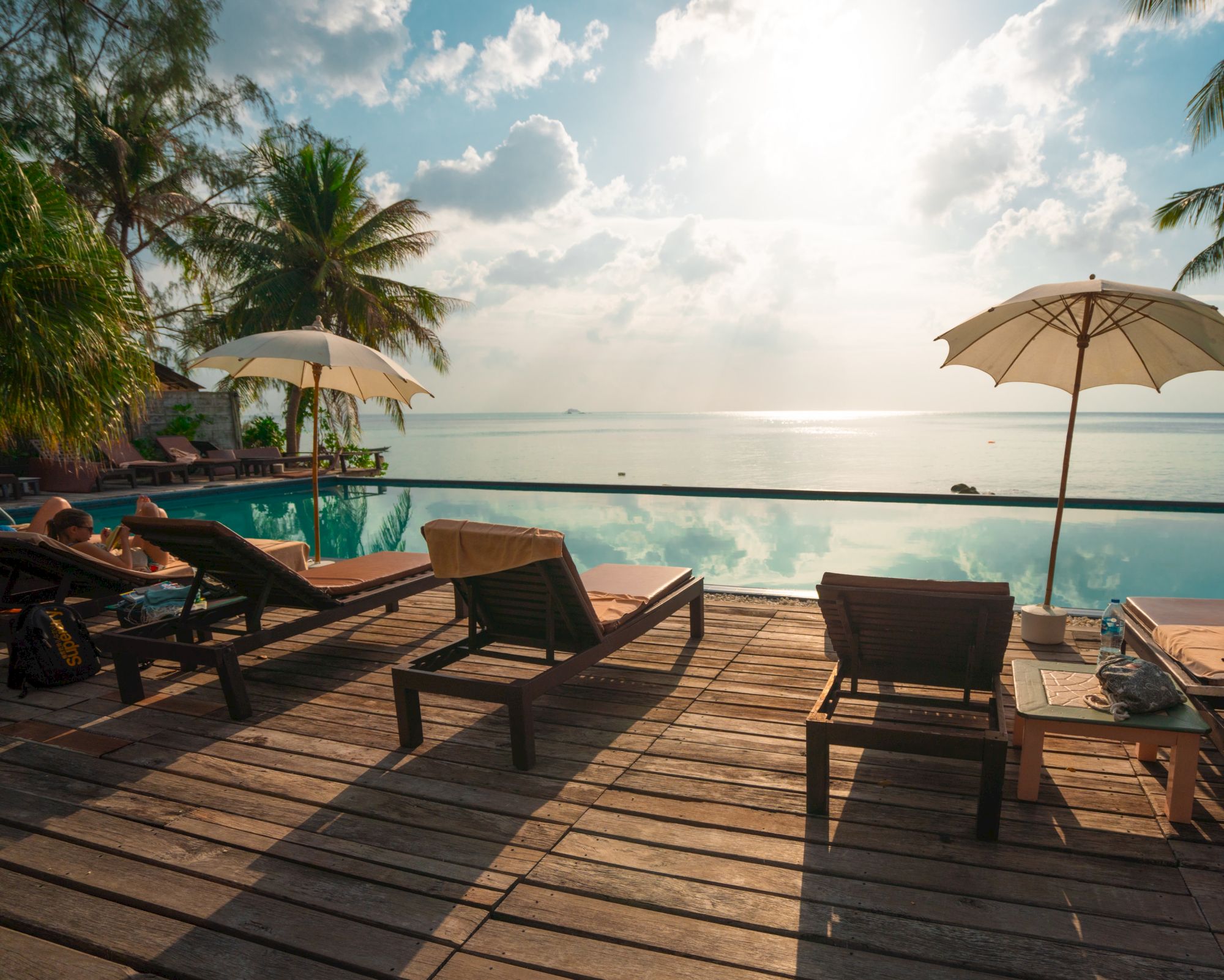 Lounge chairs and umbrellas by a pool, overlooking a calm sea with palm trees and a bright, partly cloudy sky.