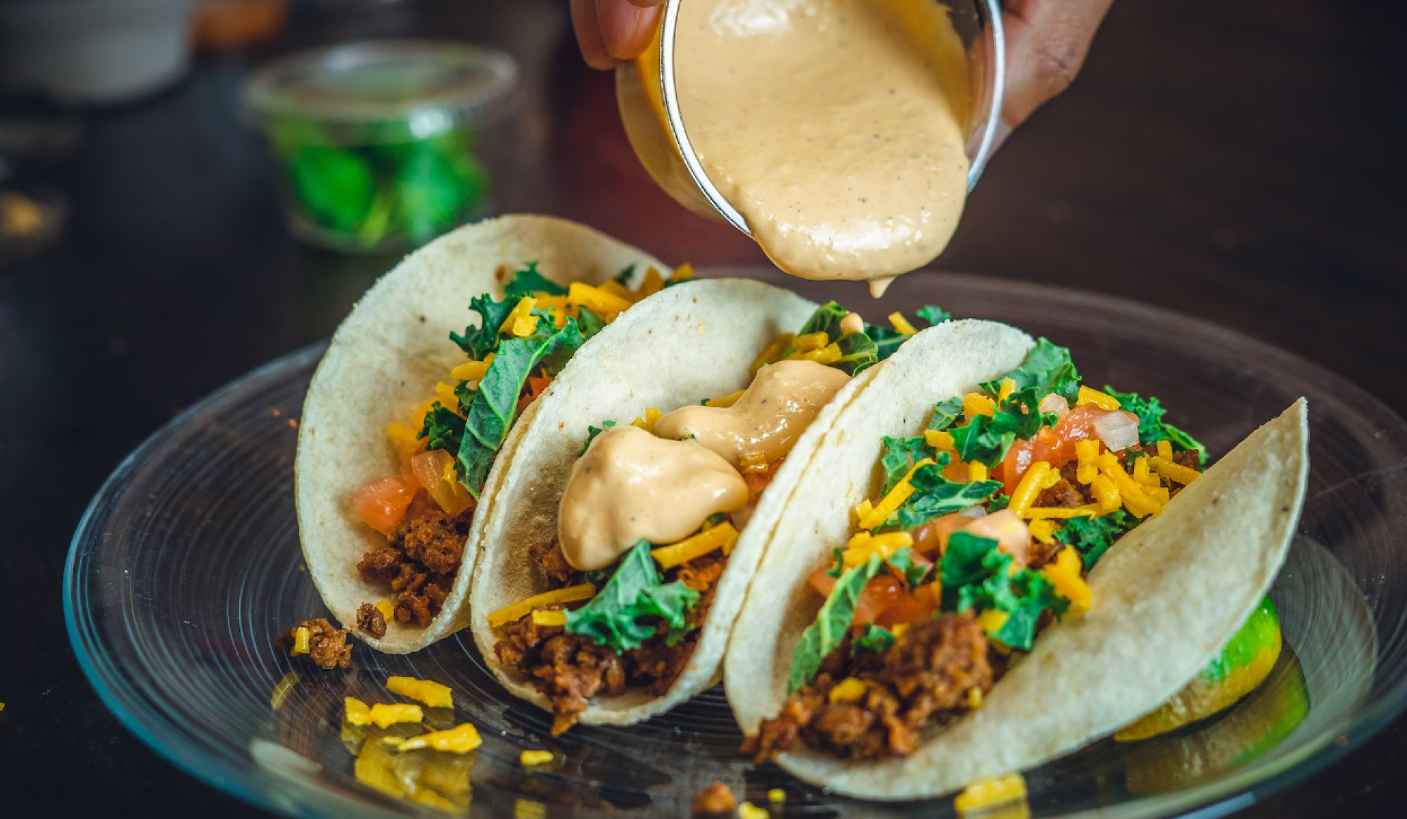 Three tacos filled with seasoned ground meat, shredded cheese, kale, tomatoes, and a creamy sauce being poured on top.