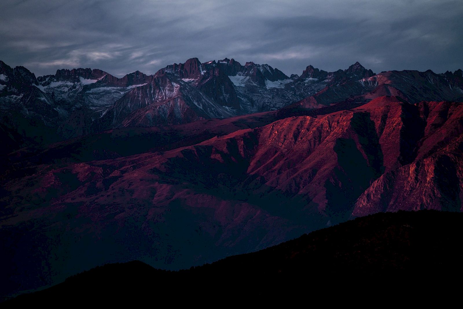A mountainous landscape is bathed in warm, red light during sunset, with jagged peaks in the background under a cloudy sky.