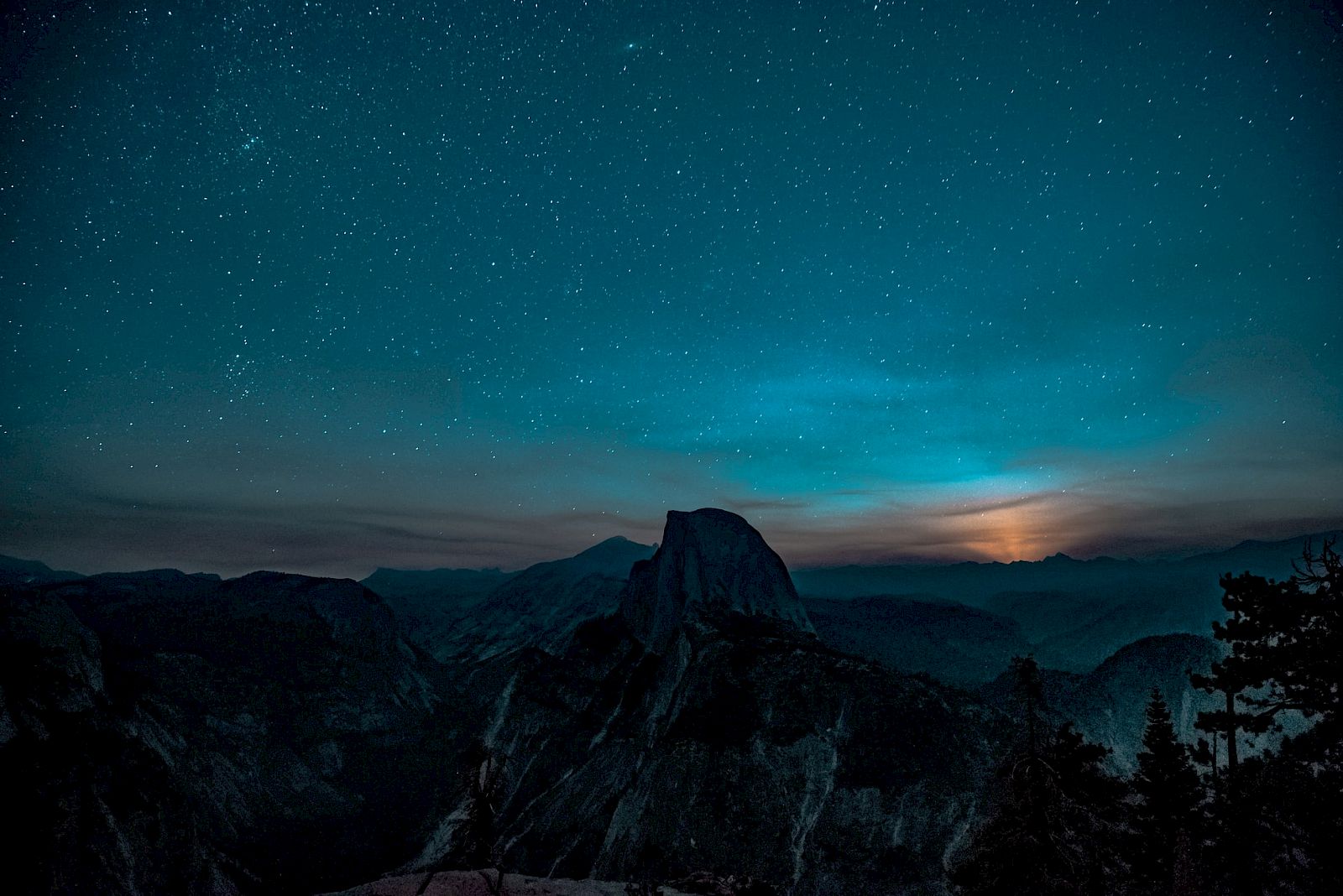 A starry night sky over a mountainous landscape, with trees silhouetted in the foreground and a bright light near the horizon.