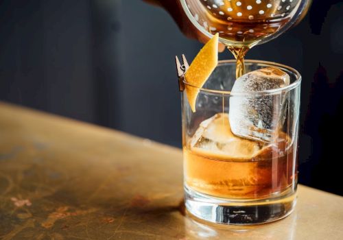 A cocktail is being poured into a glass with ice, garnished with a twist of citrus peel clipped to the rim.