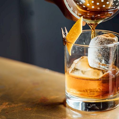 A cocktail is being poured into a glass with ice, garnished with a twist of citrus peel clipped to the rim.