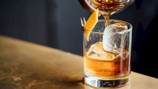 A cocktail is being poured into a glass with ice, garnished with a twist of citrus peel clipped to the rim.