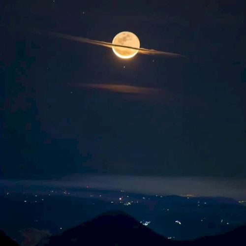 The image shows a full moon in a dark sky, with clouds giving it a ring-like appearance, resembling Saturn, above a distant, illuminated landscape.