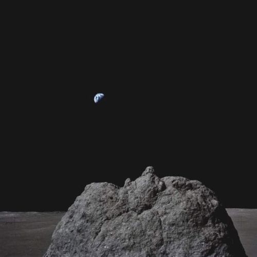 A lunar surface with a large rock in the foreground and Earth visible in the distant background against the dark sky.