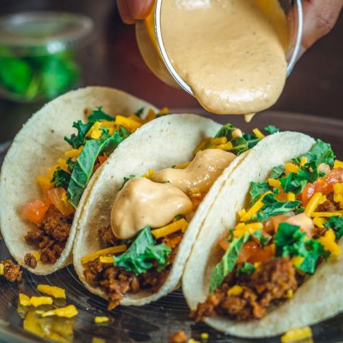 Three tacos filled with seasoned meat, cheese, tomatoes, and greens being topped with a creamy sauce on a plate.