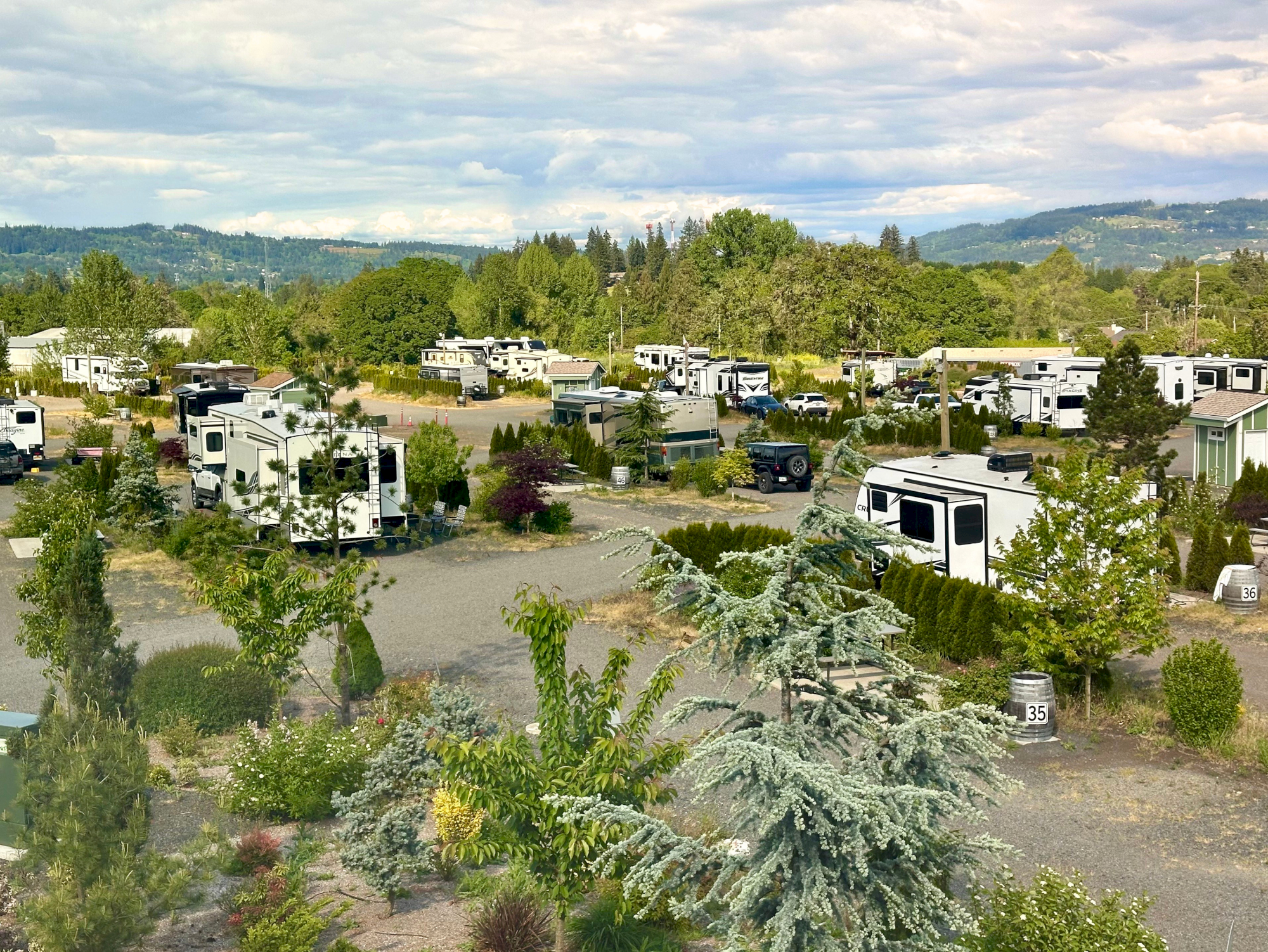 A campground packed with white RVs and camper vans, surrounded by lush trees and rolling hills under a cloudy sky.