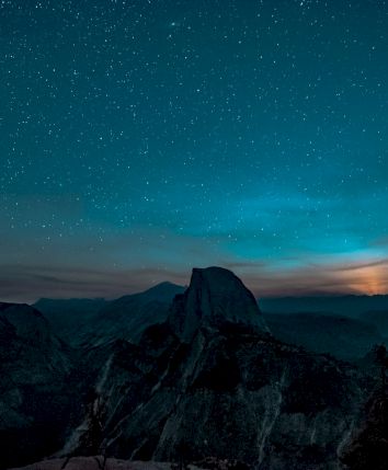 A starry night sky over a mountainous landscape with a glowing horizon and silhouettes of distant peaks and trees.