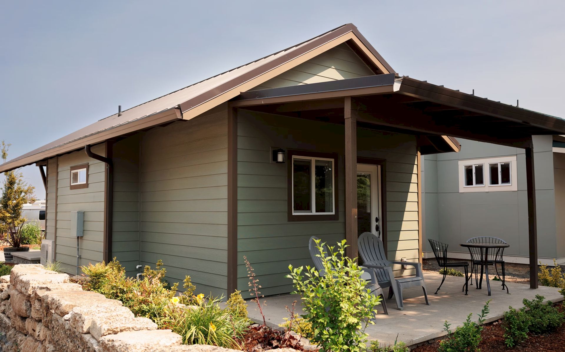 A small green house with a sloped roof, a front porch, two chairs, a table, and surrounding plants and shrubs.