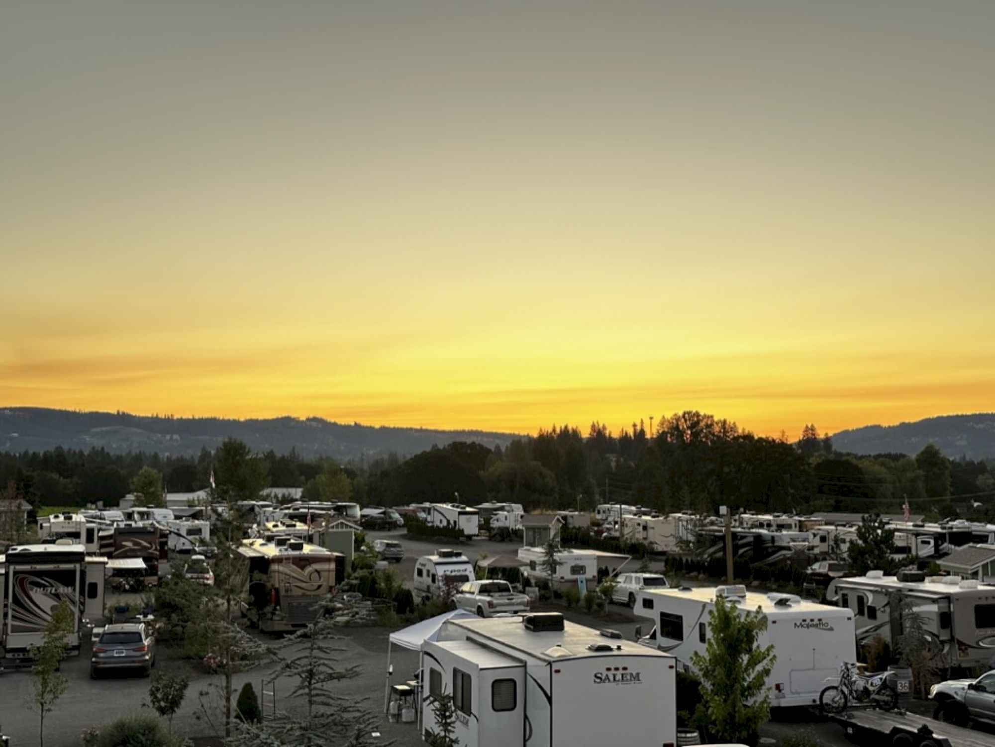 A sunset view with numerous RVs parked in a campground, surrounded by trees and hills in the distance.