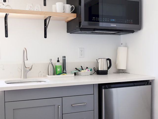 Small kitchen with a sink, gray cabinets, microwave, mini-fridge, kettle, and shelf with mugs. Paper towel roll and soap are visible.