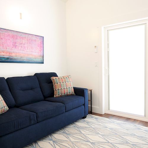 A room with a dark blue sofa, patterned pillows, a geometric rug, abstract wall art, and large sliding glass doors in the background.