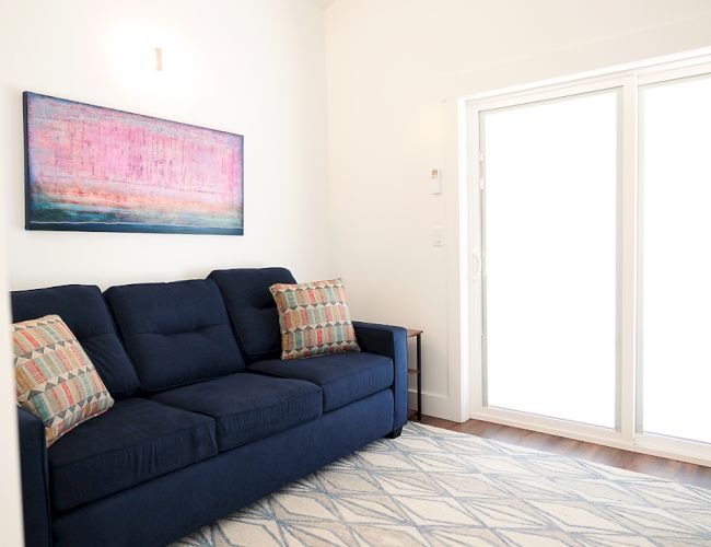 A room with a dark blue sofa, patterned pillows, a geometric rug, abstract wall art, and large sliding glass doors in the background.