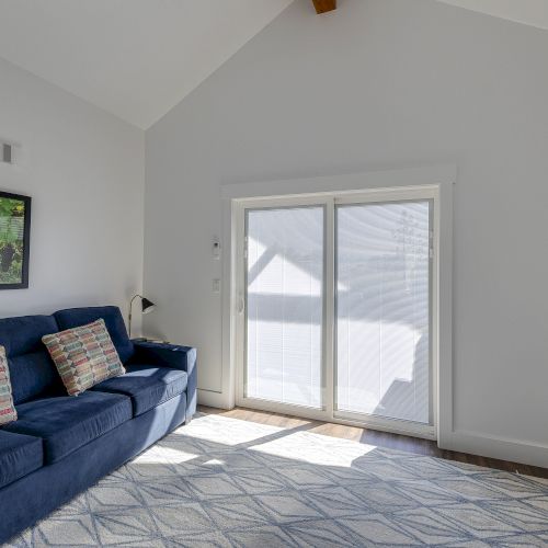Bright living room with a blue sofa, framed wall art, TV on the right, and large windows with blinds.