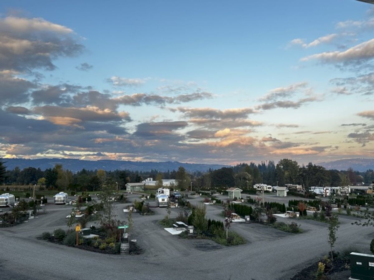 An RV park with several vehicles and trees, surrounded by a scenic landscape and cloudy sky at sunrise or sunset.