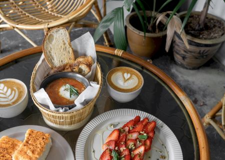 A table with two cappuccinos, bread with sauce and poached egg, mentaiko toast, and a plate of strawberries with mint leaves and pistachios.