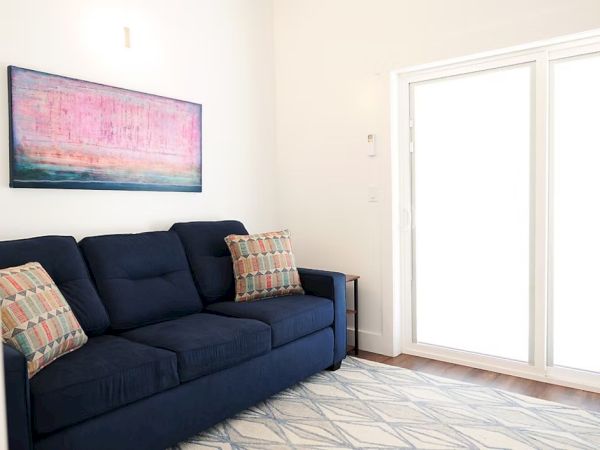 A cozy living room with a navy sofa, colorful cushions, abstract artwork, and large sliding glass doors letting in bright light.