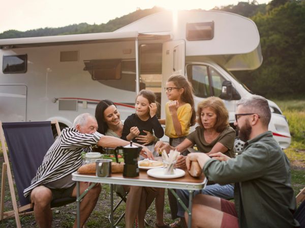 A group of people enjoy a meal outdoors by a camper van, surrounded by nature. Some are seated in chairs, with food and drinks on the table.