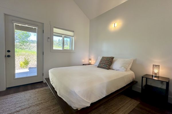 A minimalist bedroom with a bed, two lamps, a small window, and glass door, featuring simple decor and neutral tones.