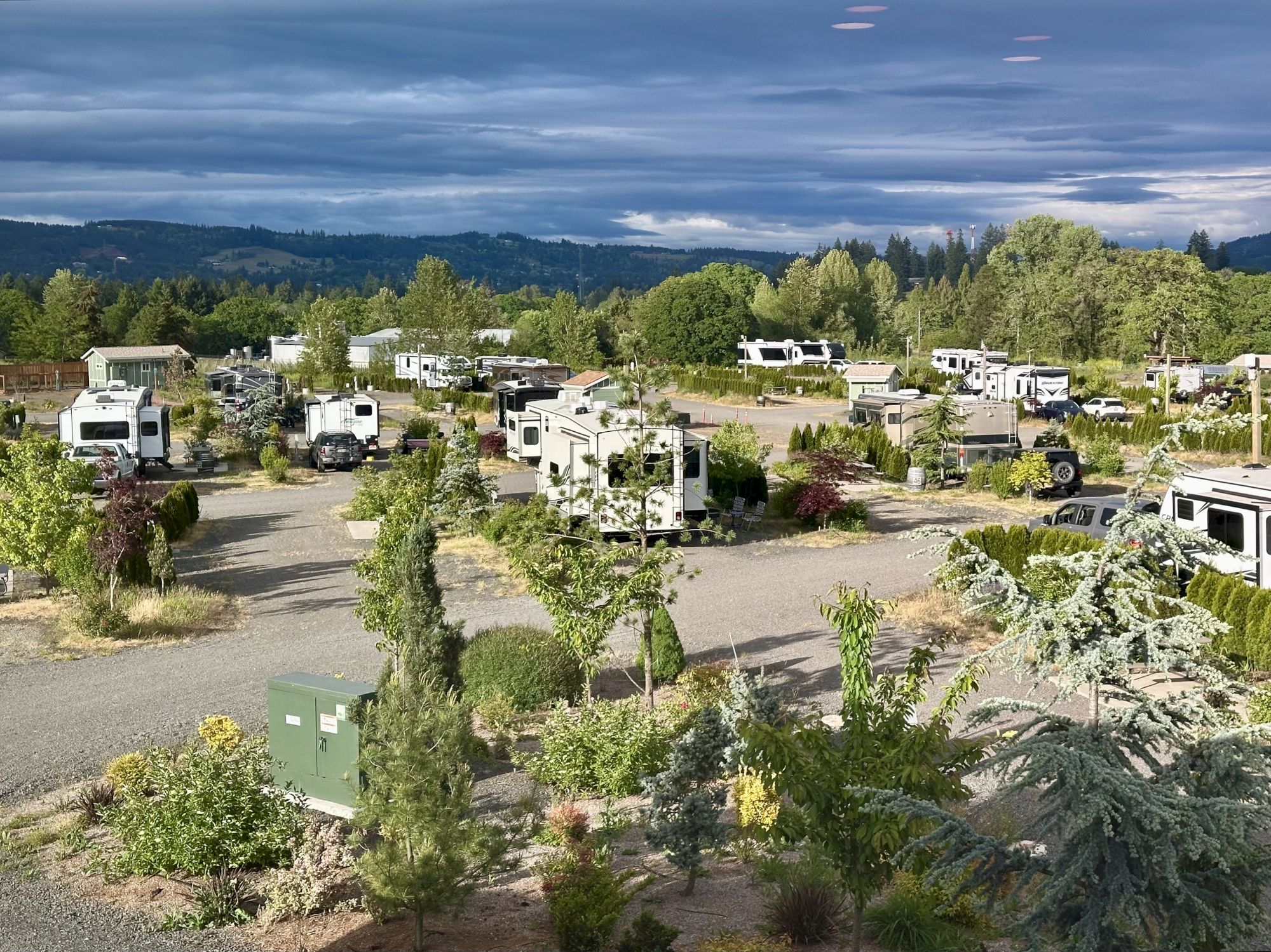 A scenic RV park with multiple RVs, trees, and greenery, set against a backdrop of rolling hills under a cloudy sky.