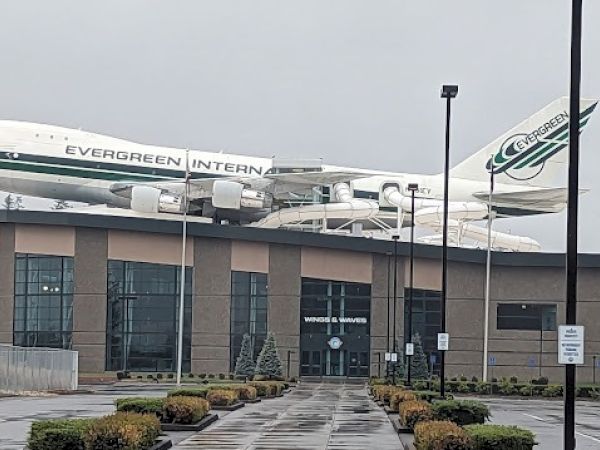A Boeing 747 with "EVERGREEN" written on it is mounted atop a building. The setting is overcast and features a parking area in front.