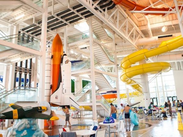 This image shows an indoor waterpark with a space shuttle model, a yellow spiral slide, and a plane model, creating a themed environment.