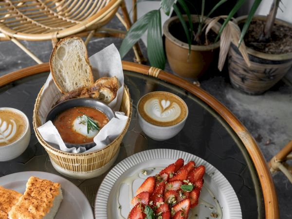 A table with coffee, bread in a basket, and a plate of strawberries, alongside potted plants and wicker chairs.