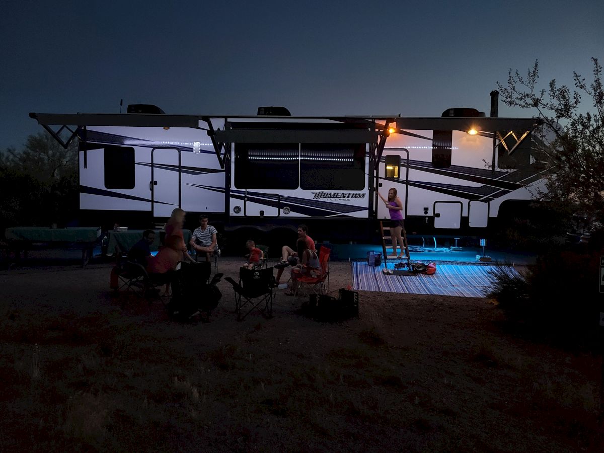 A group of people gather outdoors near a large RV, some seated in chairs under a softly lit evening sky.
