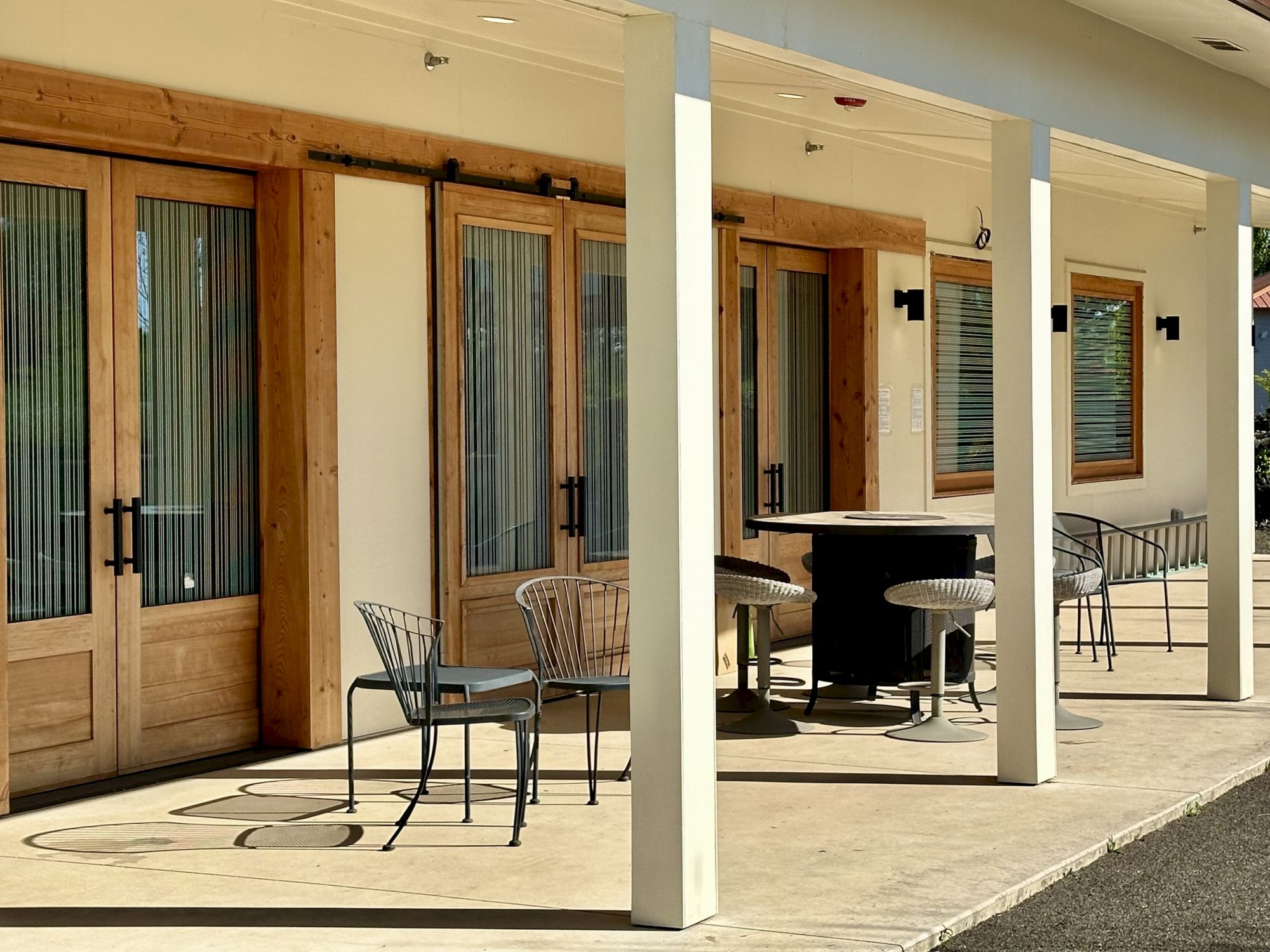 A patio with wooden doors, metal chairs, tables and windows with blinds, under a covered area with columns.