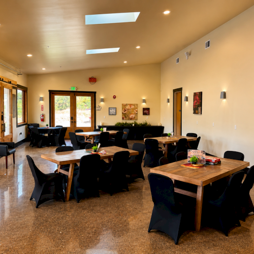 A modern community hall with wooden tables and black chairs, wall art, large doors, and warm lighting fills the room.