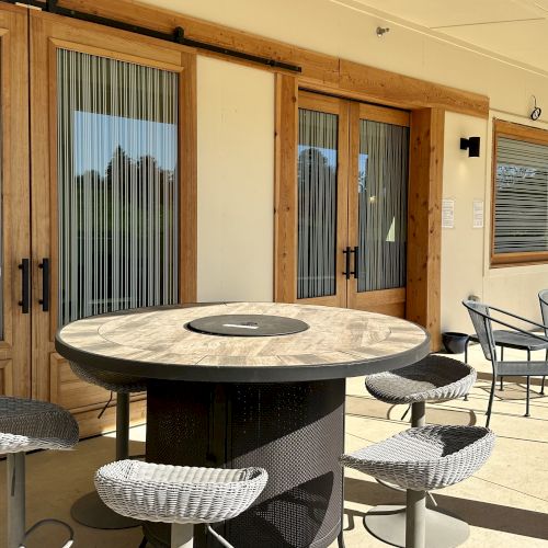 Patio with round table, four stools, wicker chairs, and glass doors with blinds. Warm, sunny atmosphere.