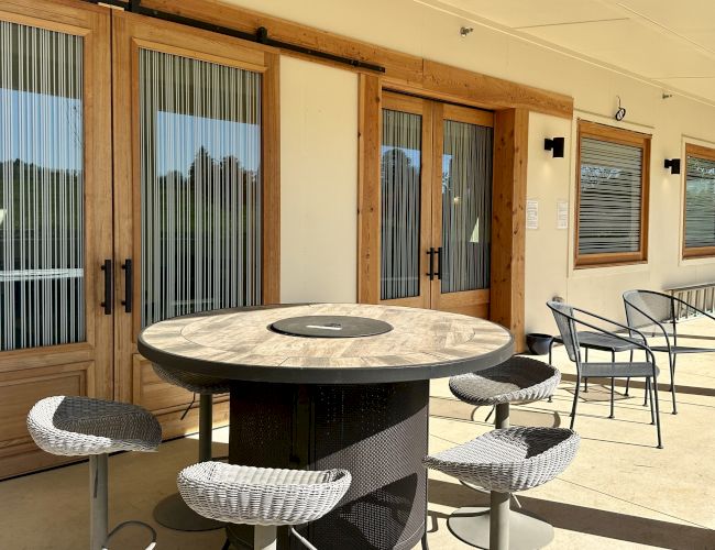 Patio with round table, four stools, wicker chairs, and glass doors with blinds. Warm, sunny atmosphere.