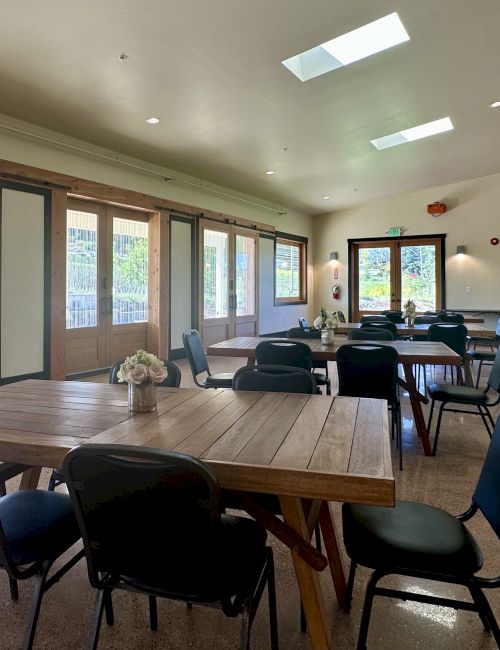 Bright room with large windows, tables, and chairs. Skylights and floral centerpieces add to the decor.